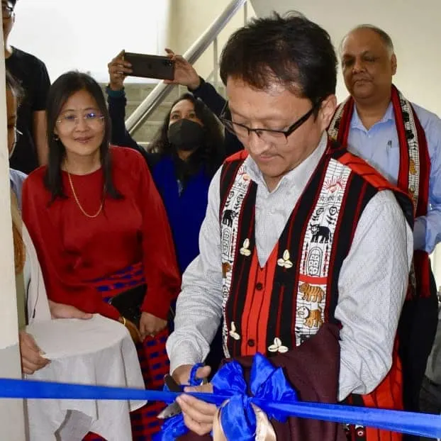 Dignitary in a traditional Naga shawl cutting a blue ribbon at an inauguration, with officials looking on