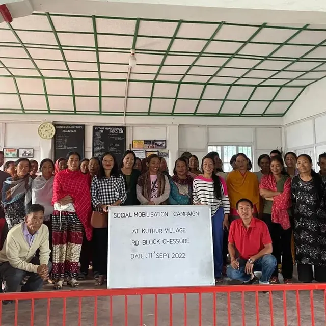 Large group of villagers seated and standing behind a Social Mobilisation Campaign notice board
