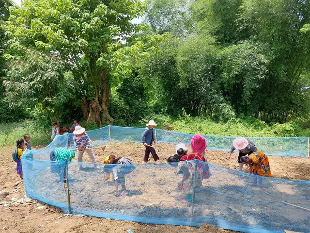 Women working in a fenced vegetable plot in a green, forested village setting