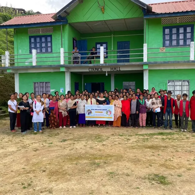 Large group photo of villagers holding a banner in front of a green two-storey building
